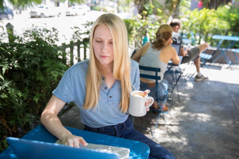A woman sits at a park, holding a mug of coffee and working on her laptop at a table