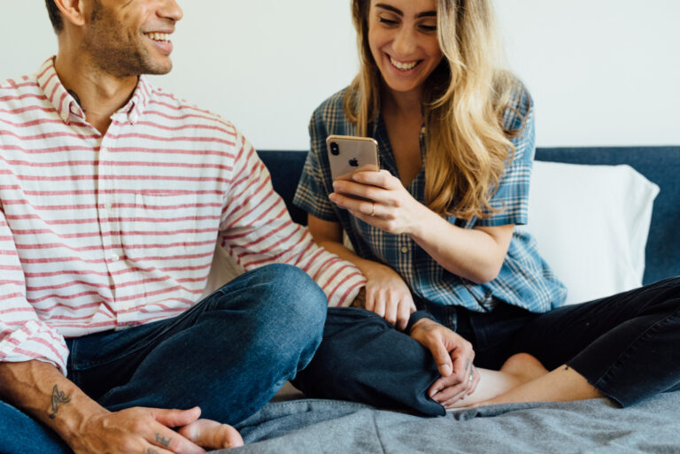 A man and woman sit cross-legged together on a bed, affectionately. The woman smiles at her phone in hand.