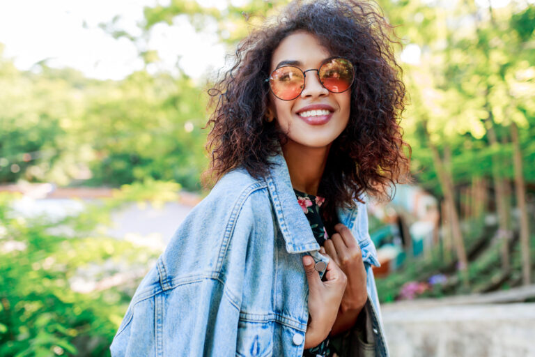 Close up of a woman smiling outdoors. She holds a denim jacket draped over her shoulders.