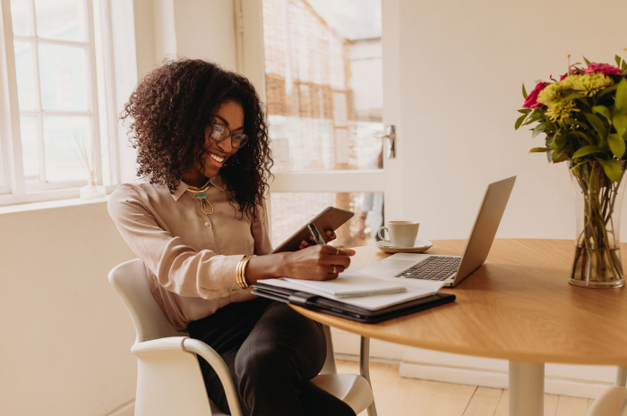 Black women sits at desk smiling and looking at her phone. There is a computer and note book on the desk in front of her.