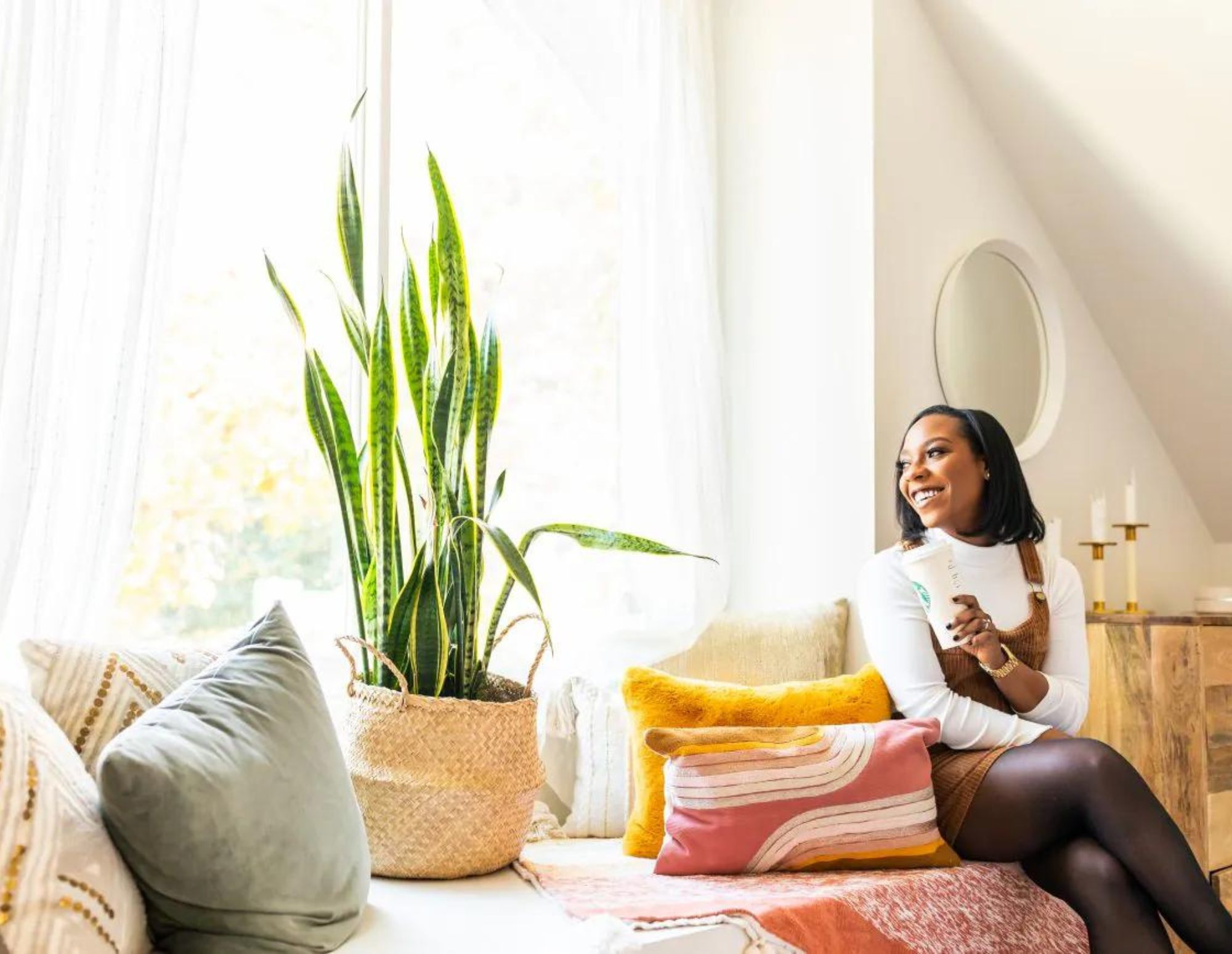 Woman gazes out of window with coffee next to snake plant.