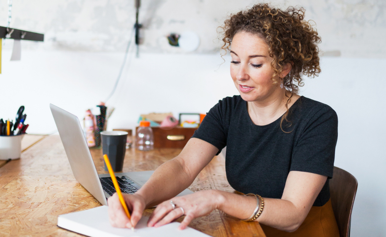 Woman sits at desk with laptop open but she's looking down at a journal and writing with a pencil