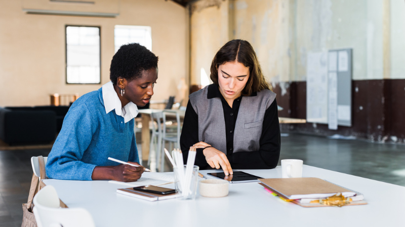 Two women site at a table looking down at an iPad during a client meeting