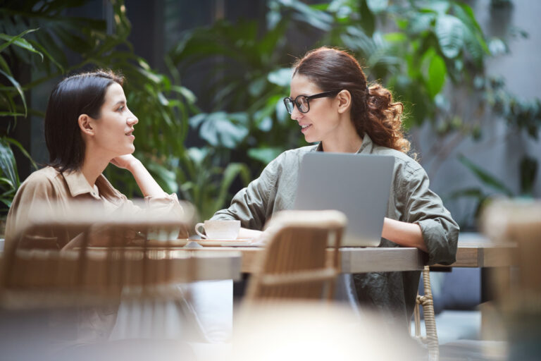 Content confident young ladies in casual shirts sitting at table in cafe and using laptop while working on web design Confident ladies working on web design