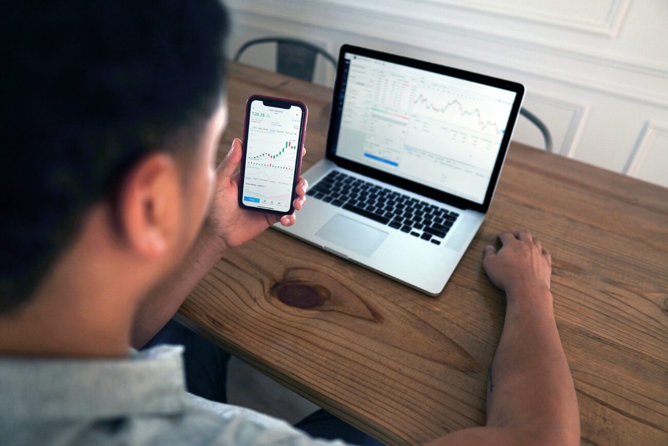 Photo shows the back of a man's head as he looks at a phone and laptop showing his freelance finances.