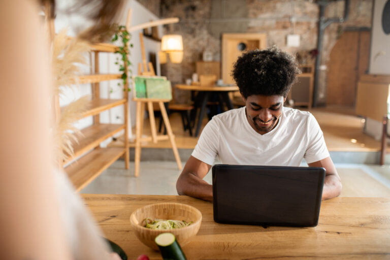 A man in a white t-shirt sits at a table, smiling down at his laptop.