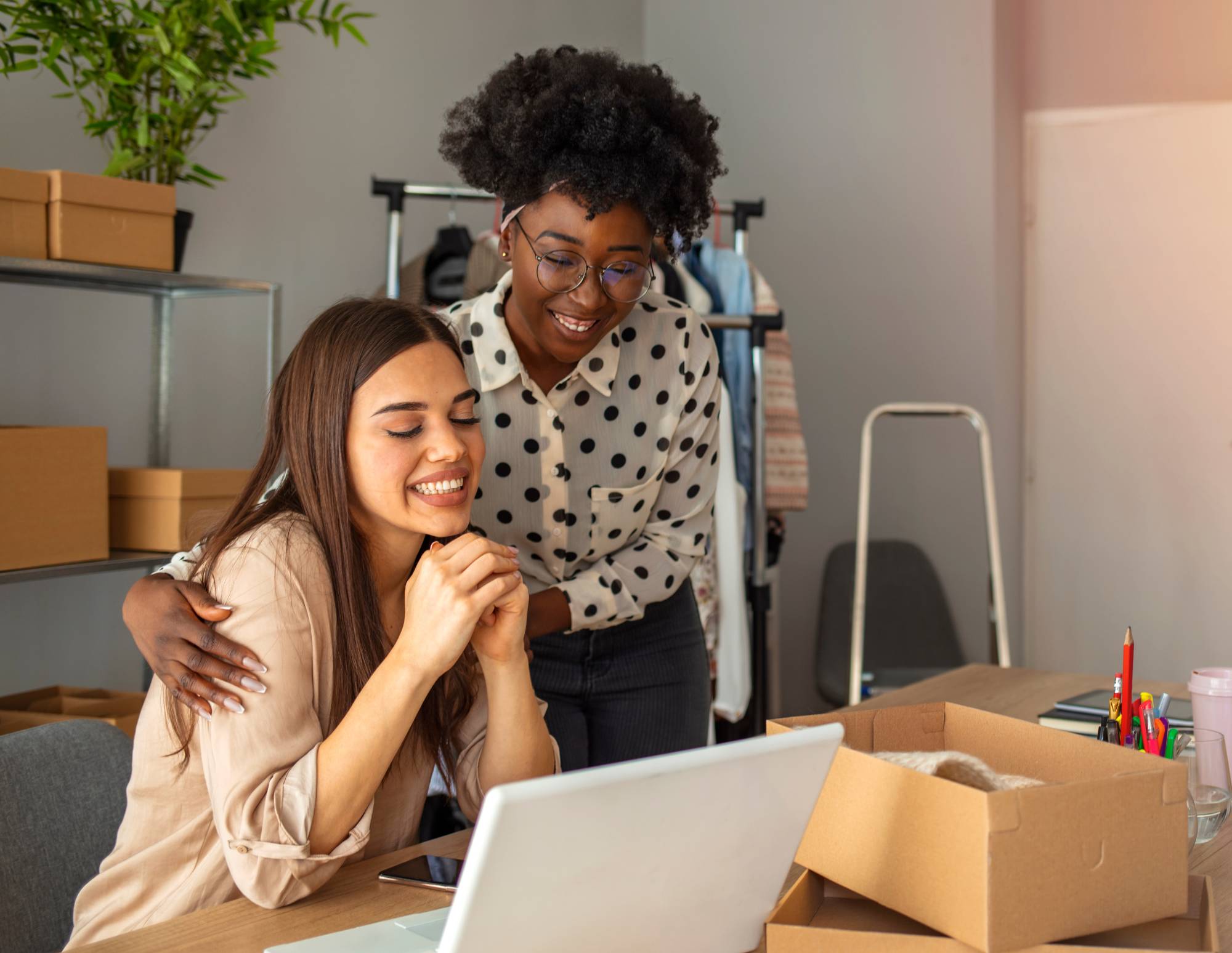 A woman sits at a desk with another woman standing beside her with arms around her shoulders. They both look happily at the computer on the desk.