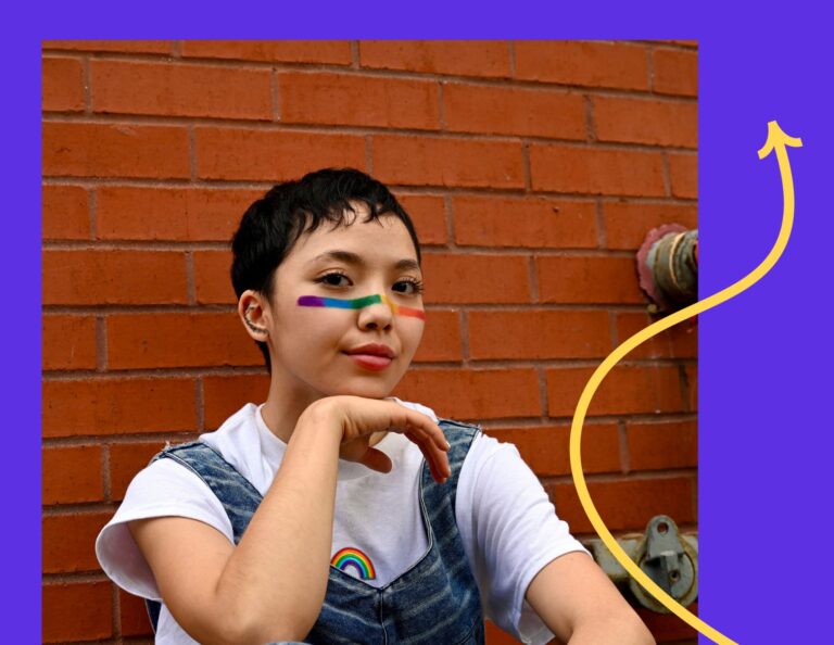 Young person leans on a brick wall celebrating Pride with rainbow facepaint.