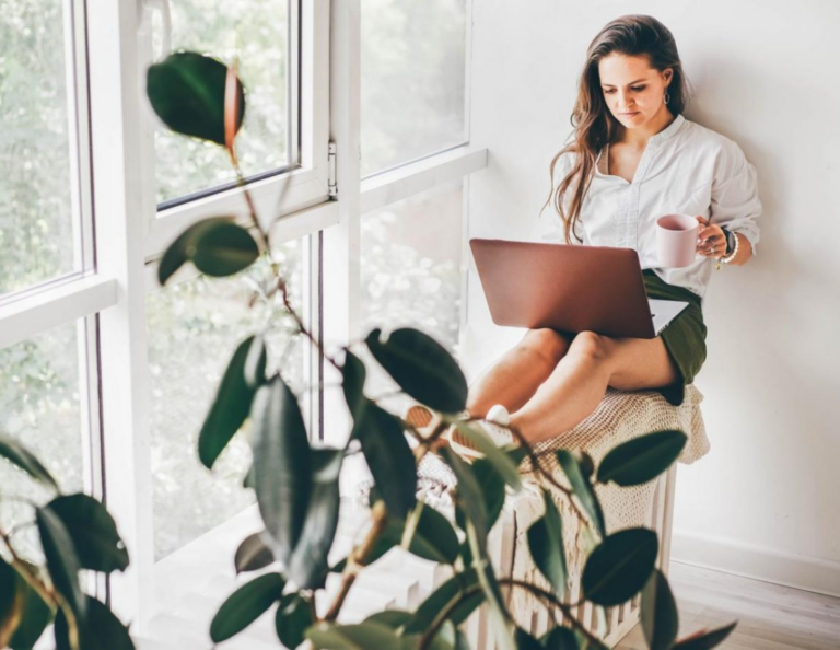 A woman sits in front of a large window, holding a mug with a laptop on her legs. There is a plant in the foreground.