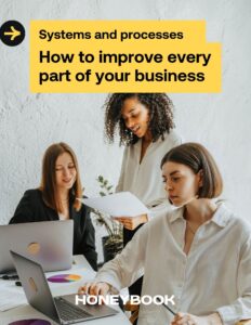 Three women look at two computers and a document on the cover of the systems and processes guide.