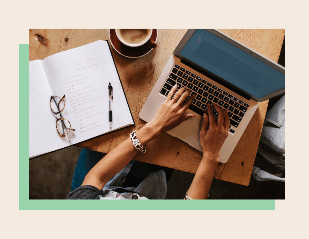 an image of hands typing on a computer with a notebook open and glasses on top of that notebook.