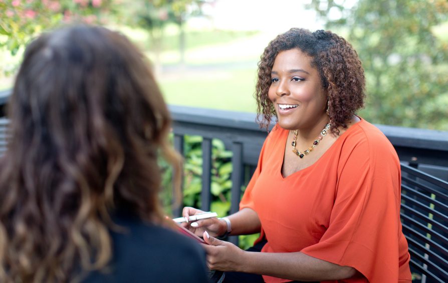 A woman in an orange shirt sits at a table outside, conversing with another person facing away from camera