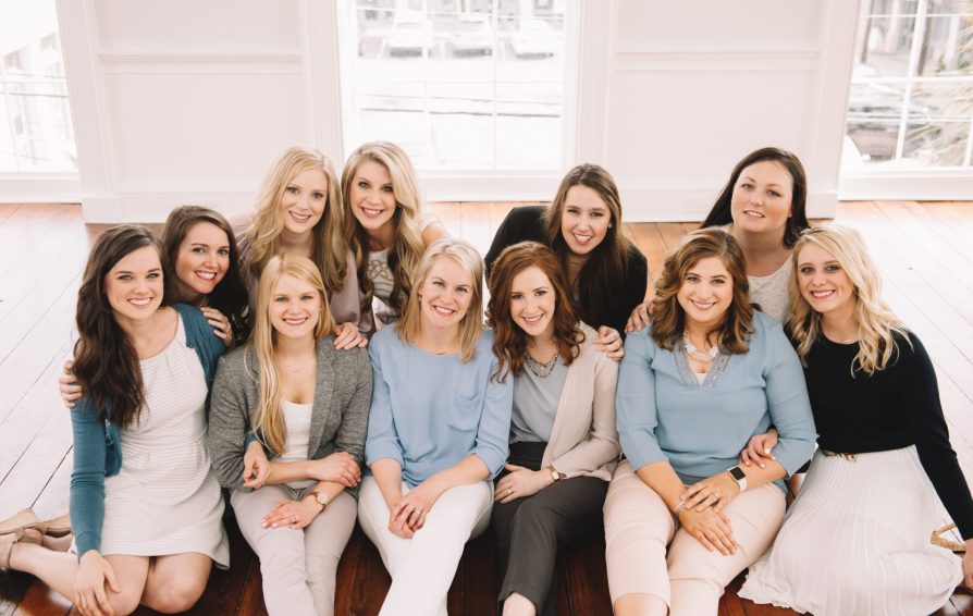 A group of women sit together on a wooden floor, smiling