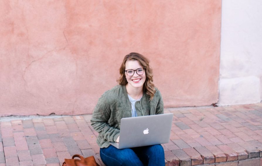 A woman sits on a brick curb, working on her laptop and smiling at the camera