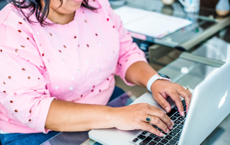 A woman in a pink sweater works at a laptop on a glass desk.