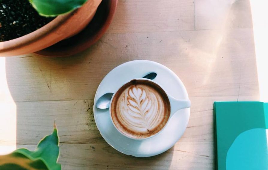 A coffee with latte art sits on a table framed by plants and assorted objects