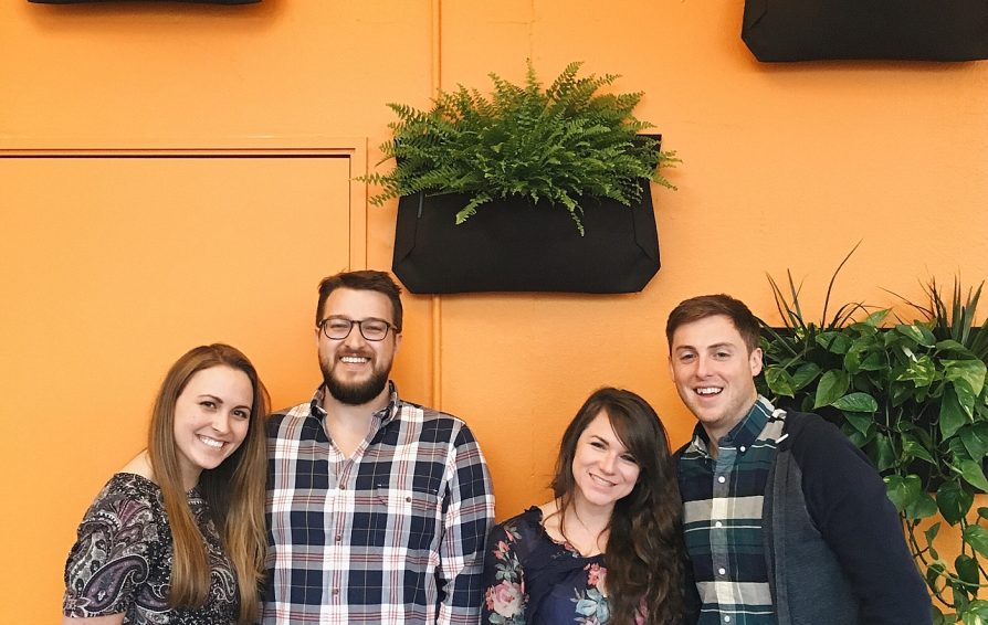 A group of people stand in front of an orange wall with plants, smiling at the camera.