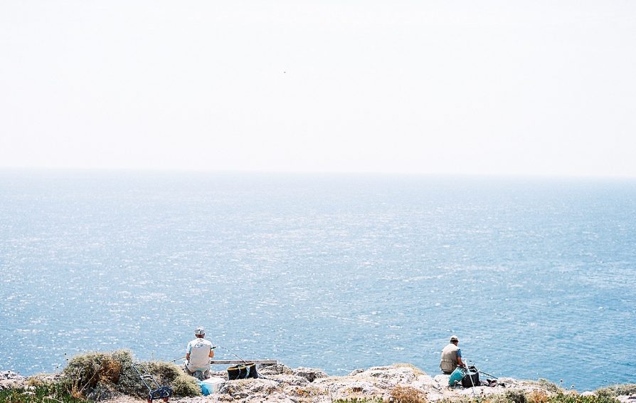 Two men sit on a cliffside, fishing into a large expanse of water.