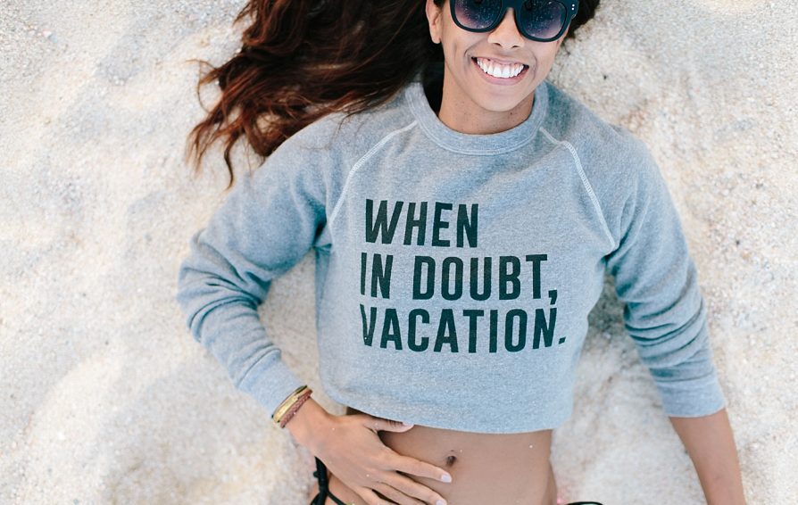 Aerial view of a woman lying in the sand, smiling. She wears sunglasses and a sweatshirt that says 