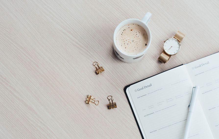 A goal planner sits on a table surrounded by a cup of coffee, watch, and binder clips