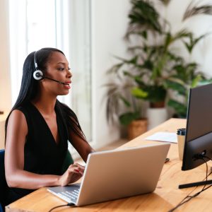 Contemporary black female with long hair wearing headset and giving instructions to client while looking at desktop monitor working at table with laptop in office.
