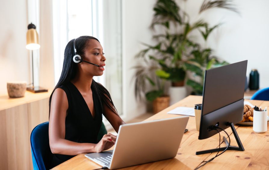Contemporary black female with long hair wearing headset and giving instructions to client while looking at desktop monitor working at table with laptop in office.