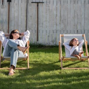 4304609 Full body relaxed mom and kid with hands behind heads looking away while relaxing on deckchairs near fence on sunny summer day in backyard.