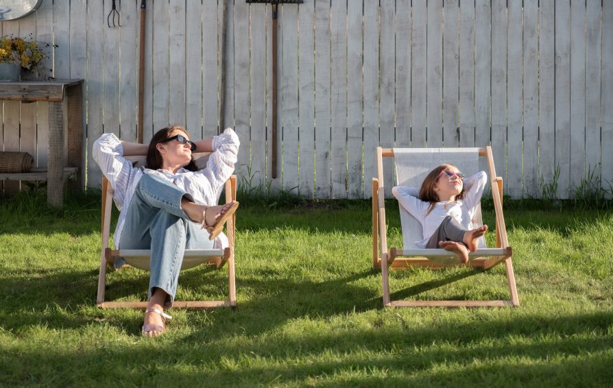 Full body relaxed mom and kid with hands behind heads looking away while relaxing on deckchairs near fence on sunny summer day in backyard.