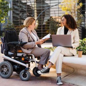 Disabled woman in wheelchair and her business colleague sitting on a park bench working on laptop and talking about business while discussing and planning new project. They are on a business meeting in front of office buildings.