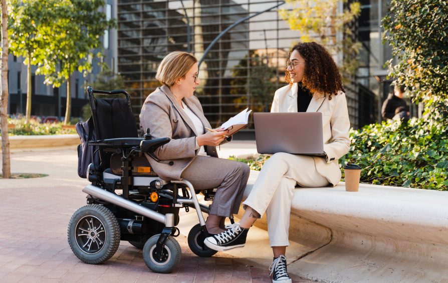 Disabled woman in wheelchair and her business colleague sitting on a park bench working on laptop and talking about business while discussing and planning new project. They are on a business meeting in front of office buildings.