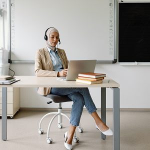 Tutor e-learning tech use device school workspace pedagogical online. Self-assured schoolteacher in headscarf conducing educational session with help of headphones and laptop from her workplace in academic environment