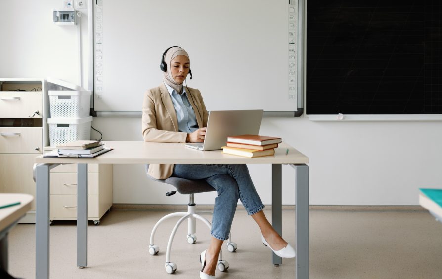 Tutor e-learning tech use device school workspace pedagogical online. Self-assured schoolteacher in headscarf conducing educational session with help of headphones and laptop from her workplace in academic environment