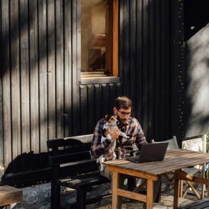Handsome young man with glasses sitting in front of his wooden cabin while holding his little white dog and working on his laptop.