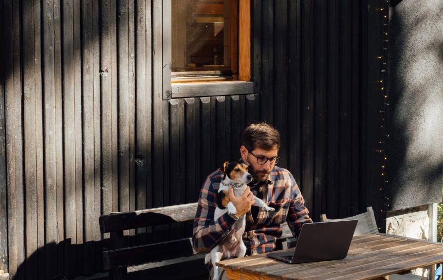 Handsome young man with glasses sitting in front of his wooden cabin while holding his little white dog and working on his laptop.