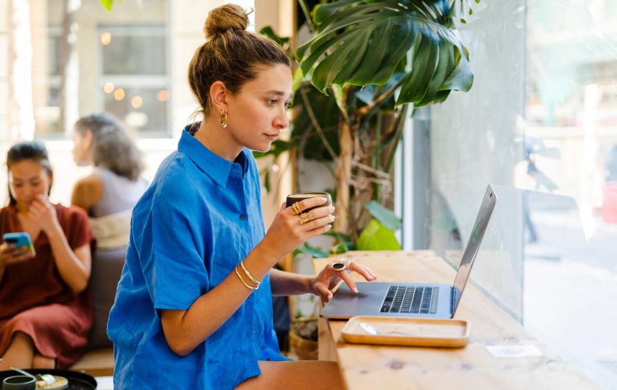 Young female entrepreneur, digital nomad, working remotely sitting in a cafe using laptop.