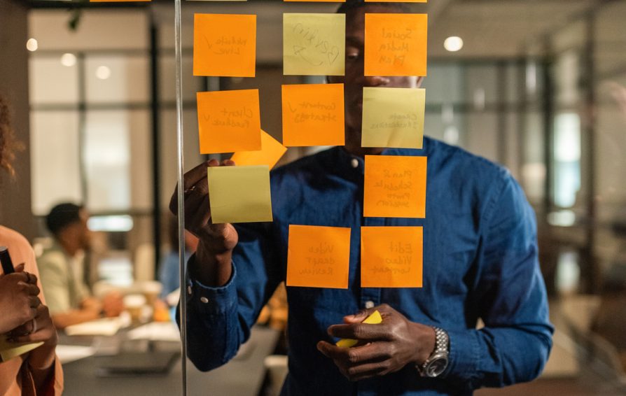 Young Black American Businessman putting sticky notes with ideas on transparent glass wall in meeting room at modern corporate office environment. Colleagues talking on background.