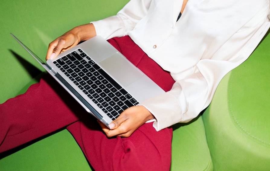 A woman working on laptop in elegant attire sits on a green chair. The modern workspace setting suggests productivity and remote work.