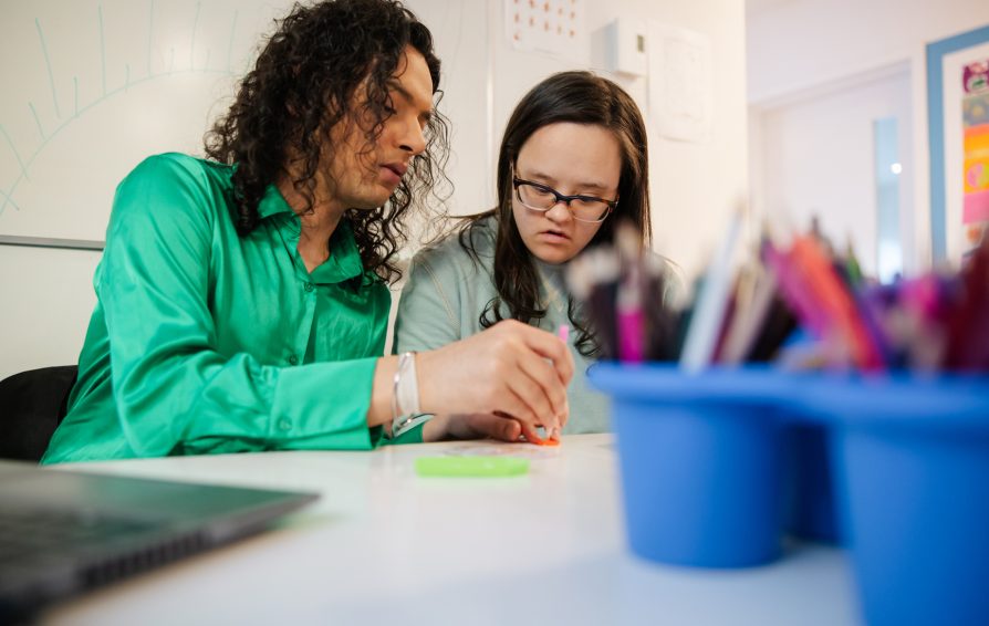 A teacher assists a student with Down syndrome in a creative learning activity at a desk. The engaging atmosphere showcases teamwork, support, and inclusion in a lively educational environment.