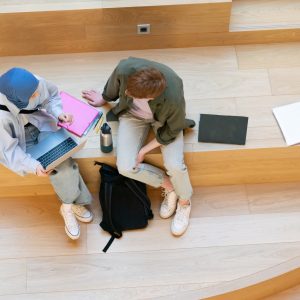 View from above of two multicultural college students sitting on wooden stairs in a bright, modern campus building, studying and chatting.