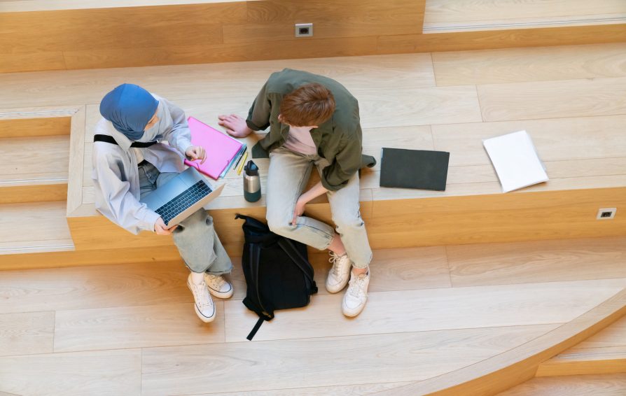 View from above of two multicultural college students sitting on wooden stairs in a bright, modern campus building, studying and chatting.