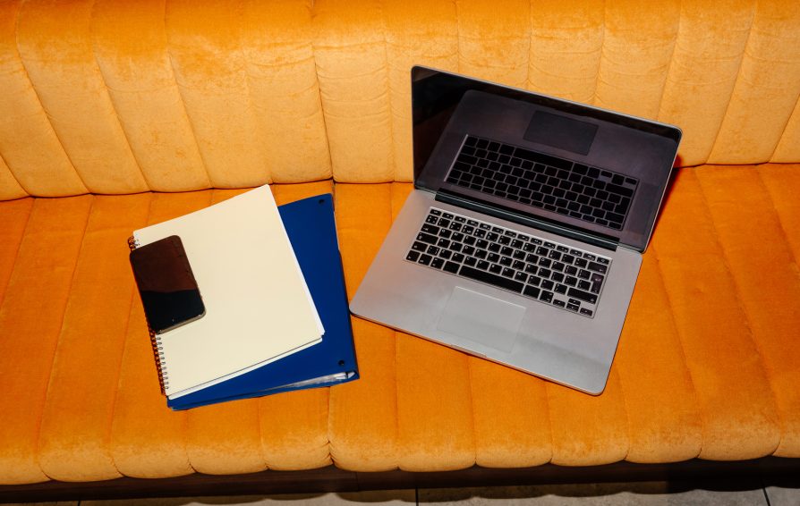 Laptop with pen, smartphone, and notepads placed on cushioned orange in modern interior