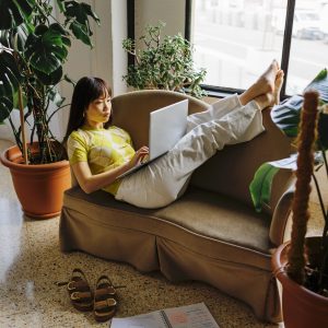 Full length of woman studying from home with laptop while lying on the couch near window in cozy living room surrounded by plants