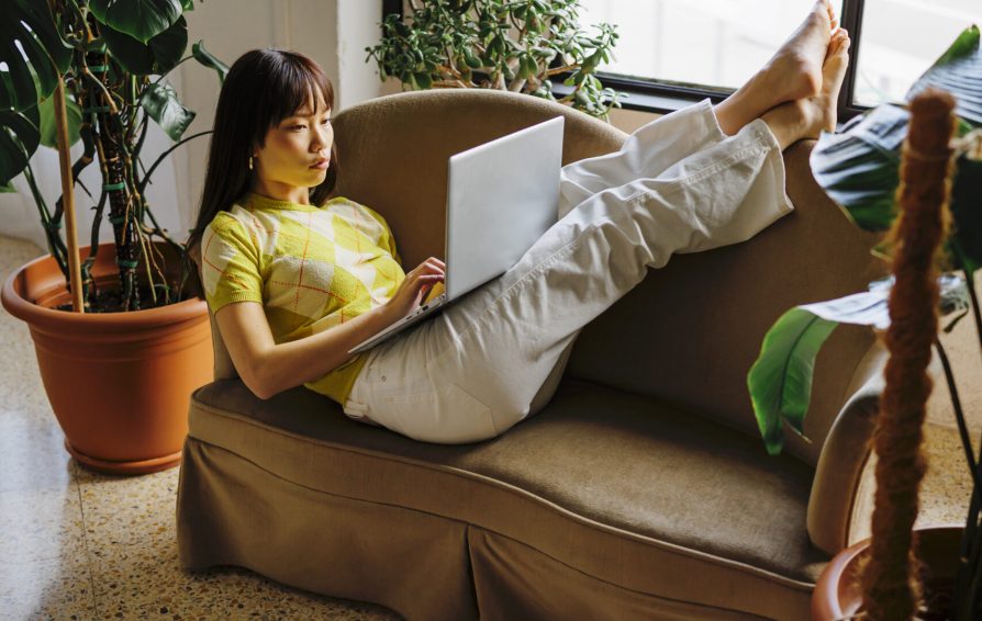 Full length of woman studying from home with laptop while lying on the couch near window in cozy living room surrounded by plants