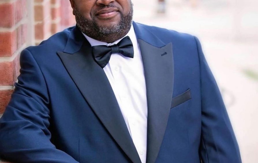 A headshot of a man in a blue suit and black bowtie, leaning against a brick wall