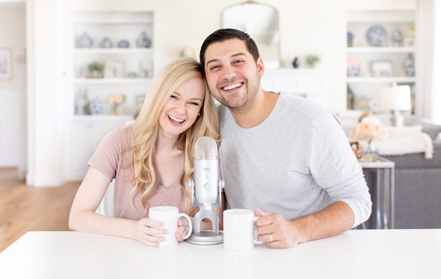 A man and woman smile with their heads together, seated behind a kitchen island. A podcasting microphone sits between them on the counter.