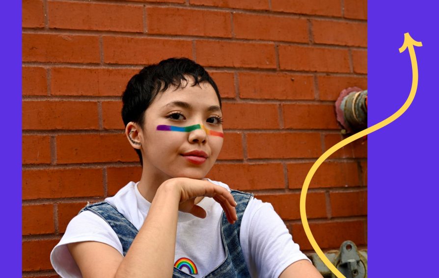 Young person leans on a brick wall celebrating Pride with rainbow facepaint.