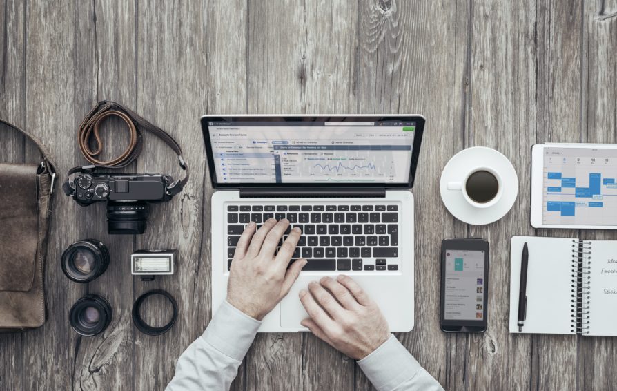 Aerial view of hands working at a laptop surrounded by camera equipment and other work supplies