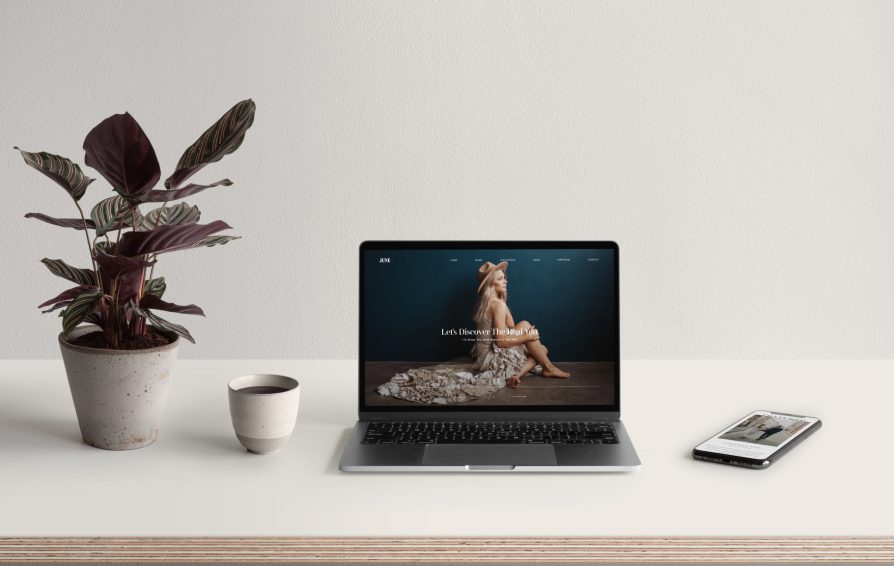 A laptop sits open with an image of a woman as the wallpaper. Beside the laptop are a cell phone, a ceramic cup, and a potted plant.