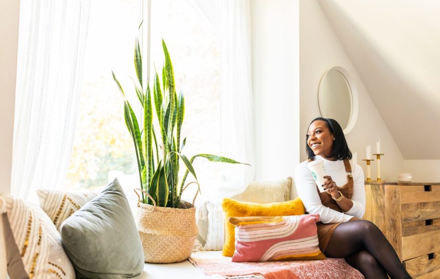 woman sitting on a couch by a large window with plants and blankets