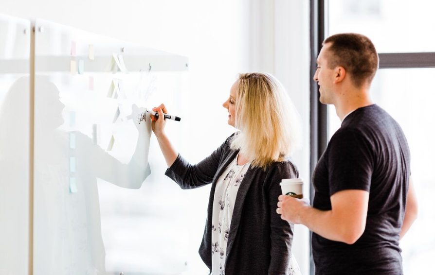 A woman draws on a whiteboard while a man holding a coffee looks on